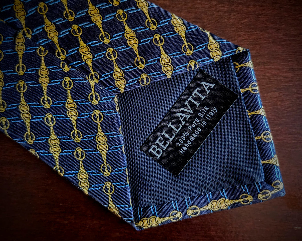 A close-up of a folded necktie on a dark wooden surface, featuring a blue and gold hanger pattern. The inner neck label clearly displays the brand 'Bellavita'.