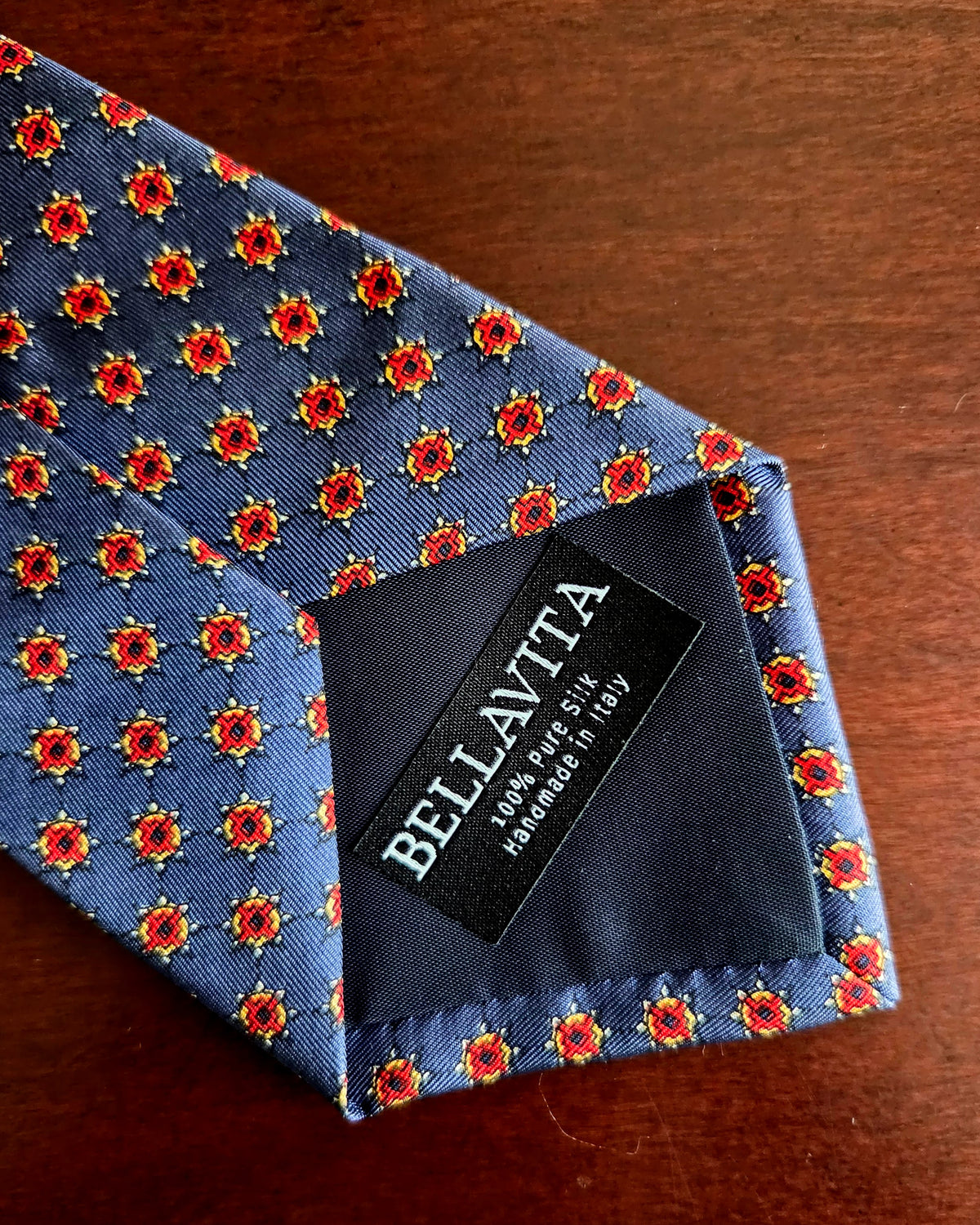 A close-up of a Bellavita men's necktie with a blue base, featuring a repeating red and gold floral pattern, laid on a dark brown wooden surface. The black fabric neckband with the white 'BELLAVITA' brand name is visible at the base.
