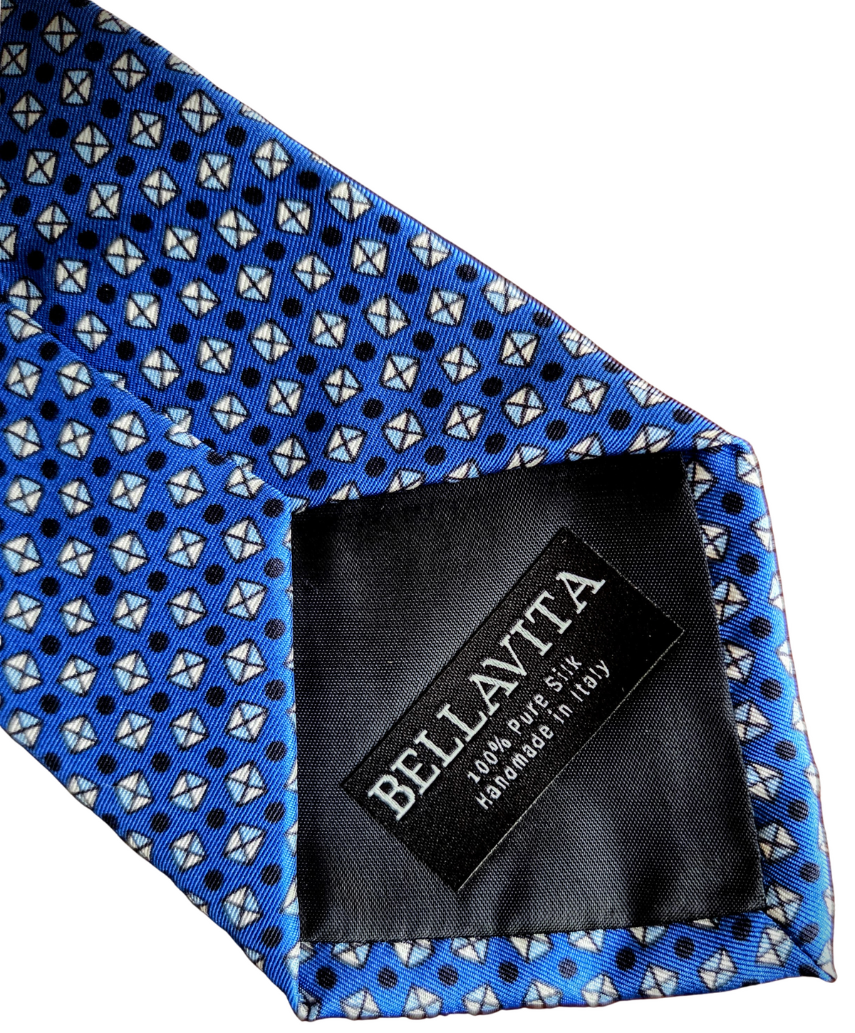 A close-up of a blue necktie with a repeating geometric pattern of white and black shapes, featuring a black fabric brand label sewn onto the inside that reads 'BELLA VITA'.