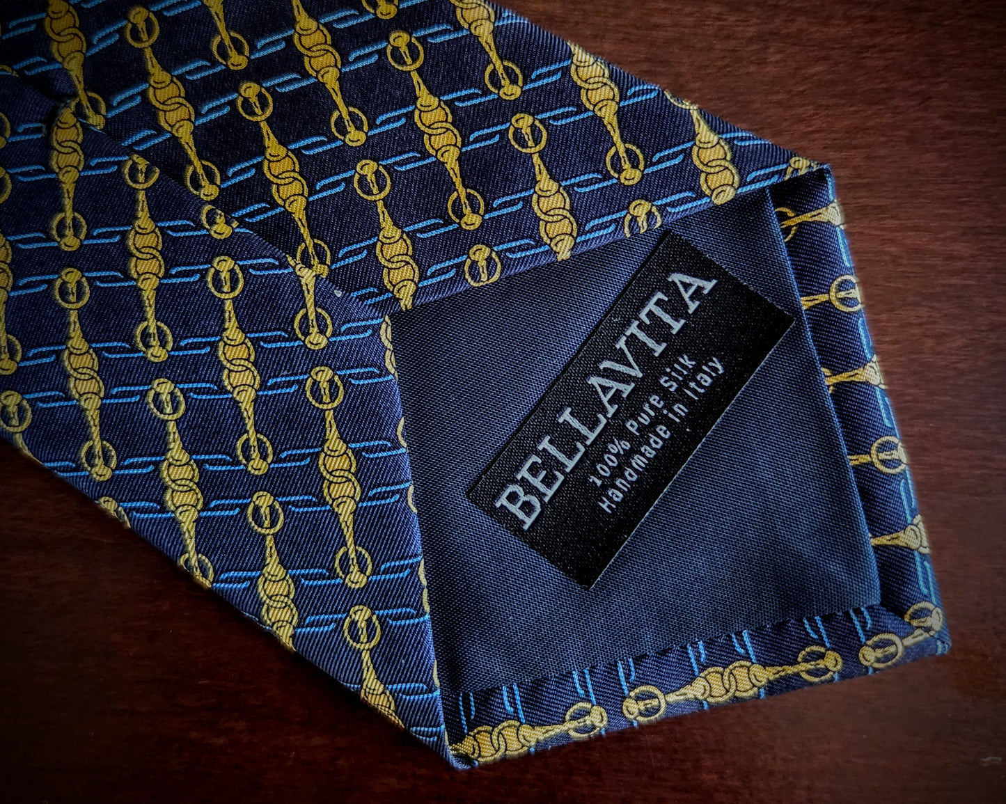 A close-up of a folded necktie on a dark wooden surface, featuring a blue and gold hanger pattern. The inner neck label clearly displays the brand 'Bellavita'.