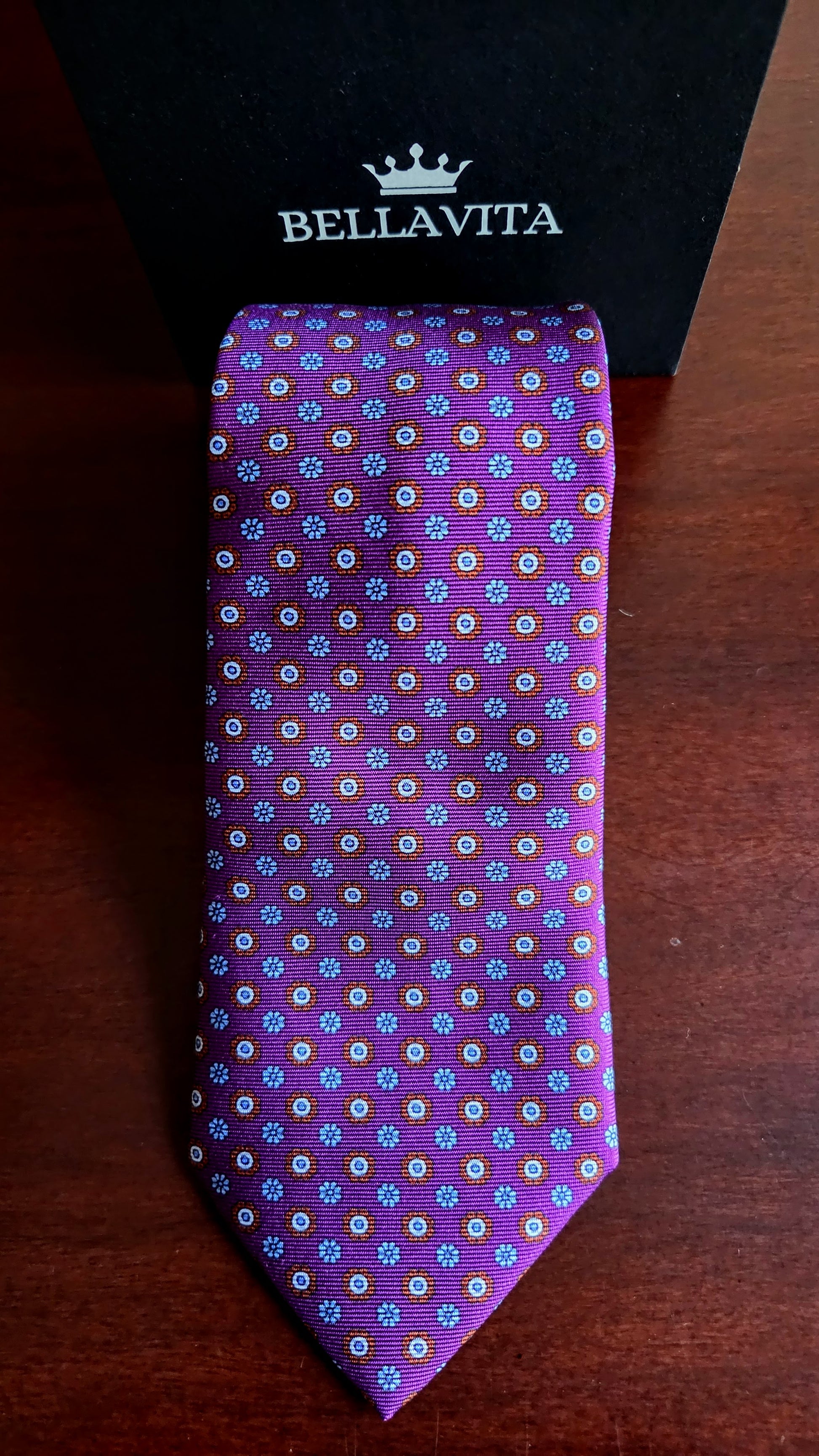 A purple silk necktie with a small multicolor floral pattern, displayed in front of its black Bellavita gift box on a dark wooden surface.
