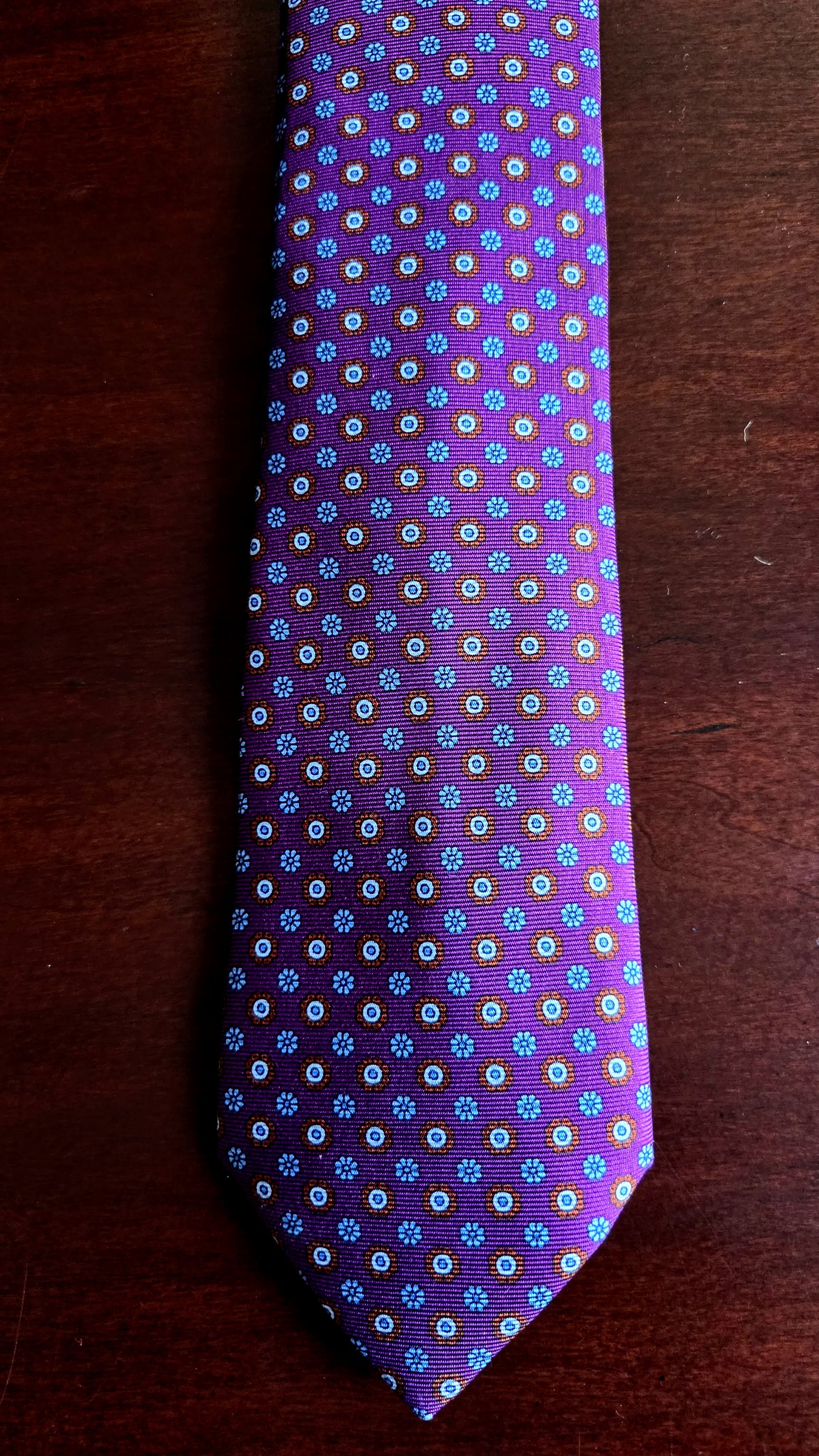 A purple necktie with a repeating small floral pattern in blue, white, and yellow, laid flat on a dark brown wooden surface.