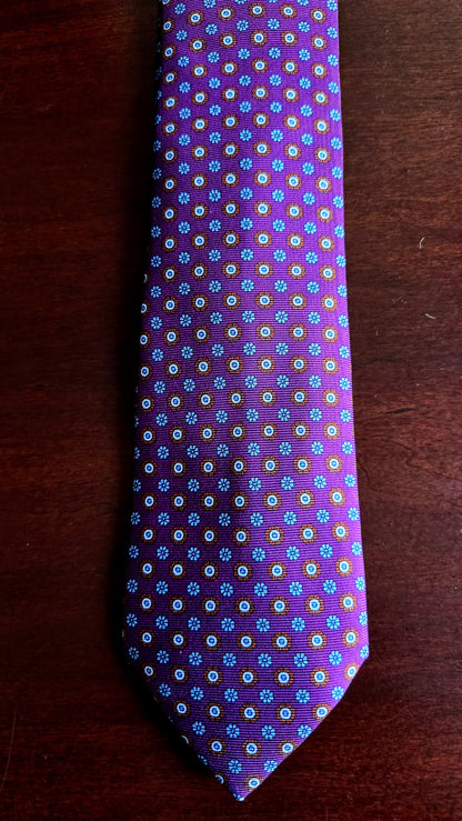 A purple necktie with a repeating small floral pattern in blue, white, and yellow, laid flat on a dark brown wooden surface.