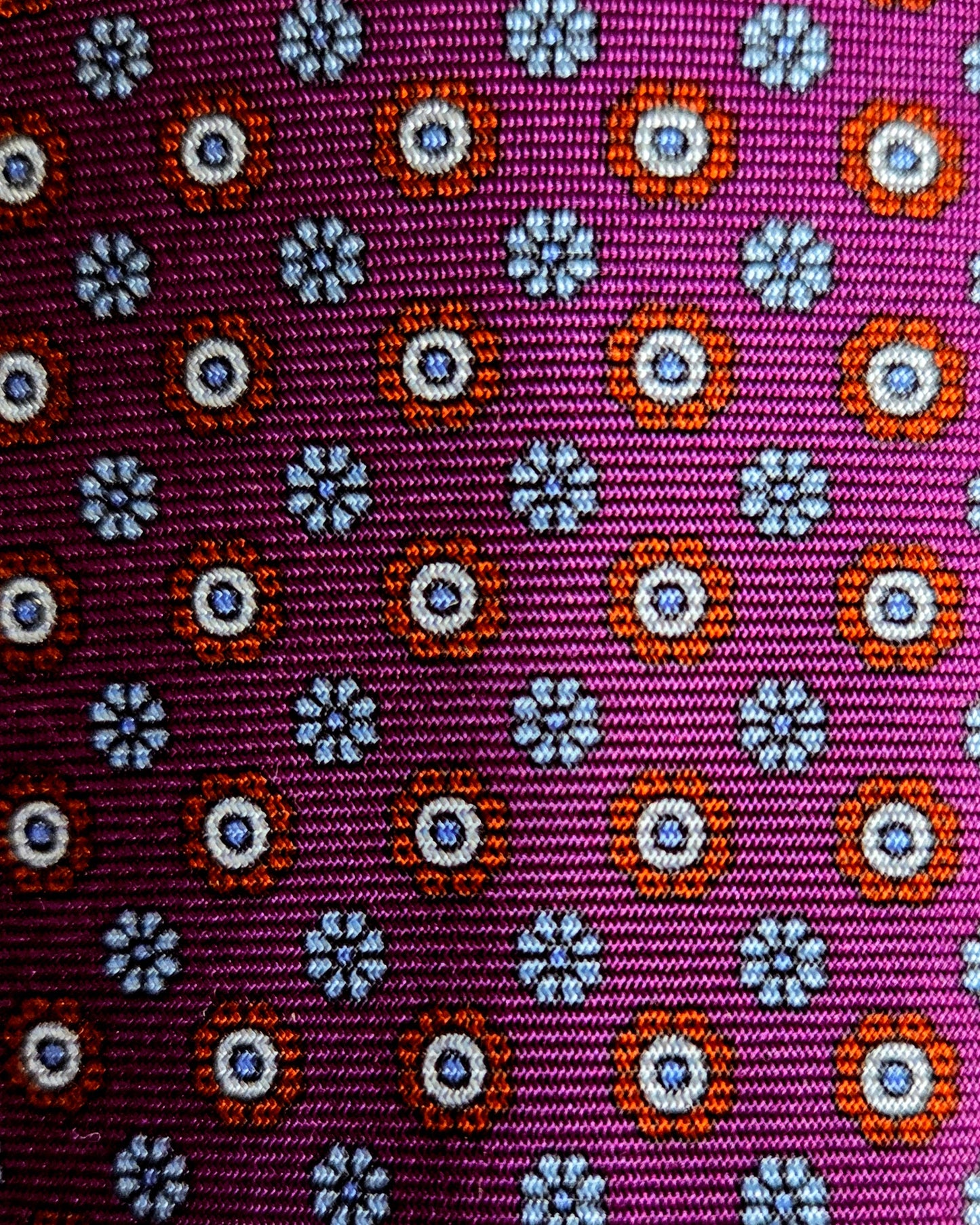 A close-up of a woven fabric with a repeating geometric pattern on a magenta-purple background, featuring orange, white, and blue circular and floral motifs.