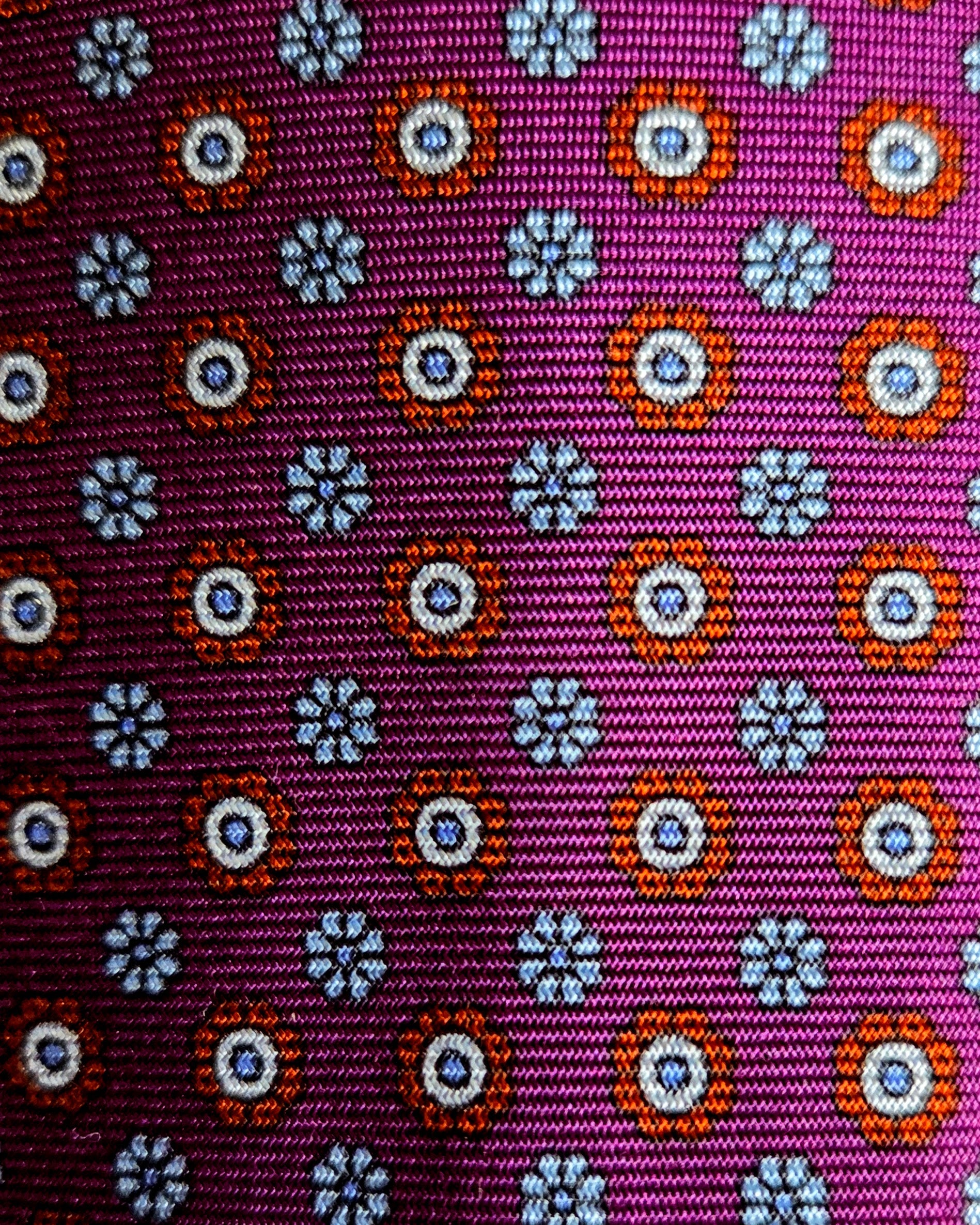 A close-up of a woven fabric with a repeating geometric pattern on a magenta-purple background, featuring orange, white, and blue circular and floral motifs.