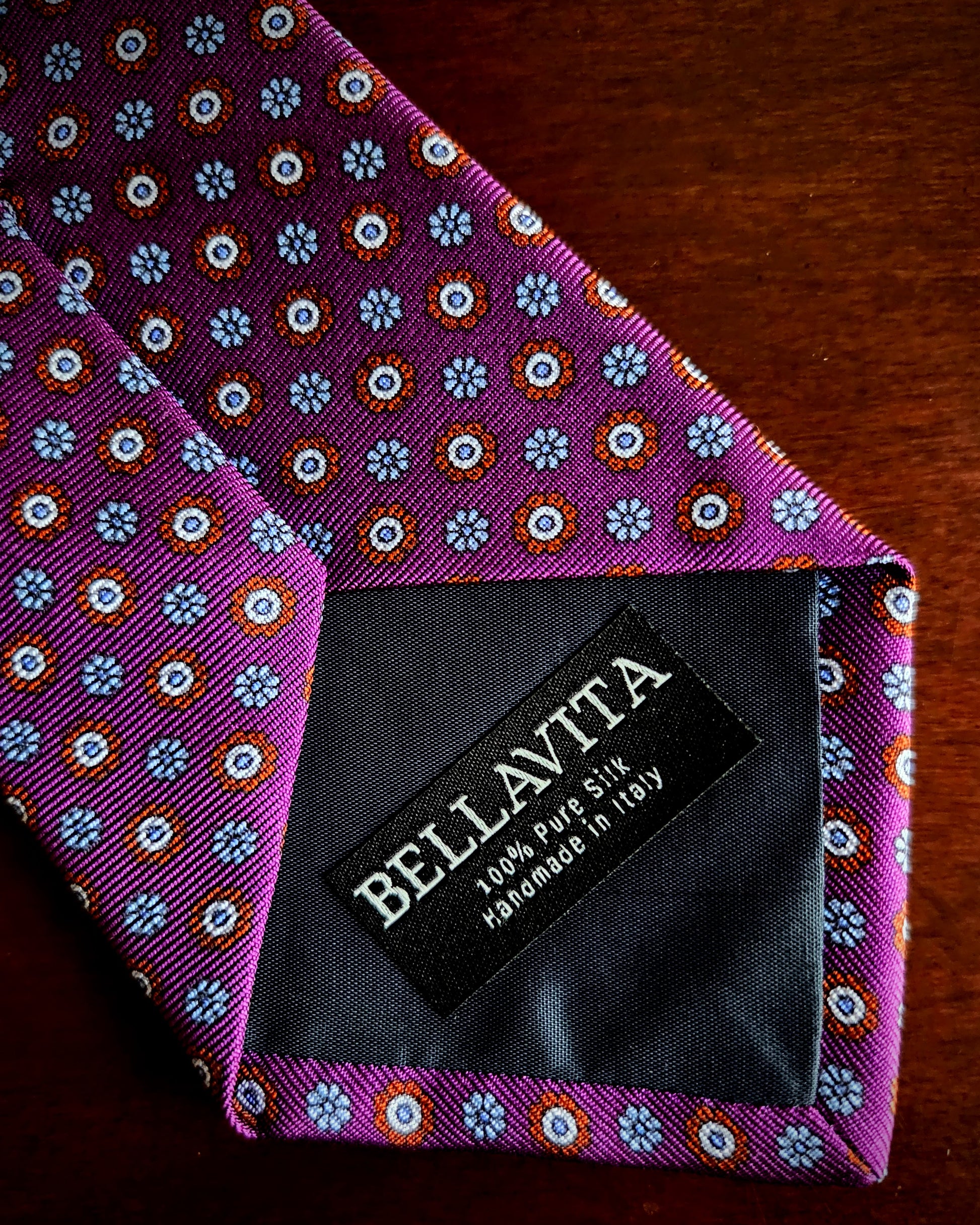 A close-up of a purple silk necktie with a repeating blue, white, and orange floral pattern, featuring a black fabric brand label that reads 'BELLAVITA 100% PURE SILK handmade in Italy'.