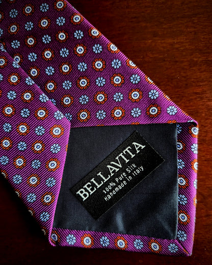 A close-up of a purple silk necktie with a repeating blue, white, and orange floral pattern, featuring a black fabric brand label that reads 'BELLAVITA 100% PURE SILK handmade in Italy'.