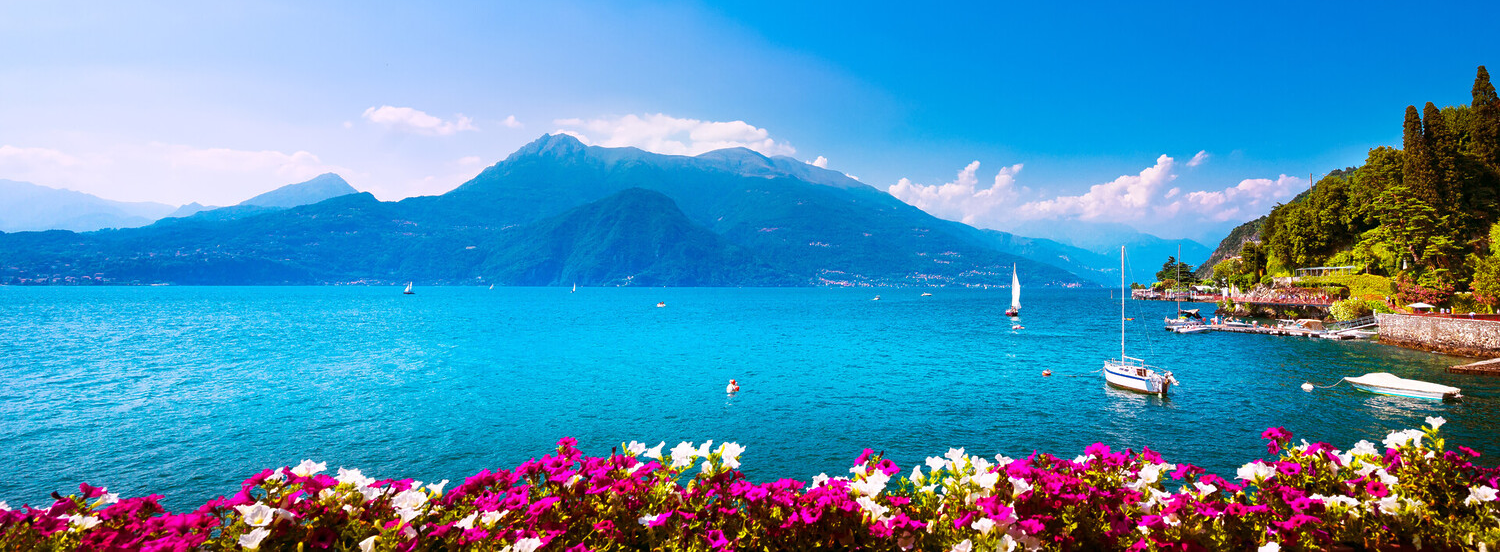 A vibrant scenic landscape print of a mountain lake, featuring bright turquoise water, sailboats, a snow-capped mountain in the background, and a foreground of colorful pink and white flowers.