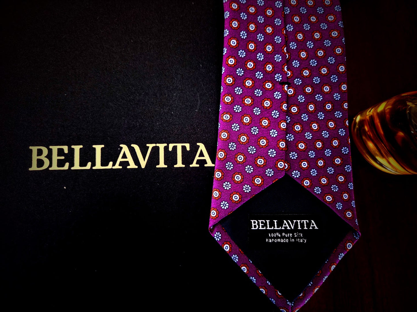 A Bellavita necktie with a purple base and a repeating multicolor floral pattern, displayed next to its black packaging box with gold lettering.